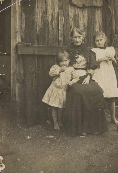 Black and white photograph of an old woman in a dark skirt sitting in front of a wooden fence, a bare-footed girl standing either side of her.