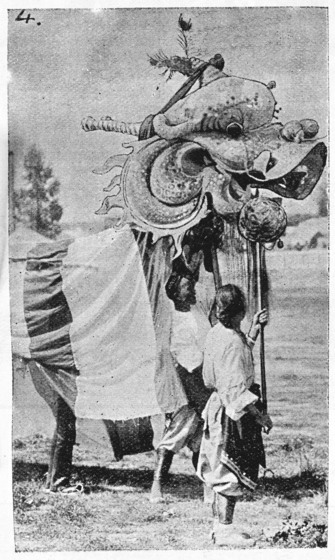 Black and white photograph of a two people supporting a large processional dragon's head with another holding a ball on a pole.