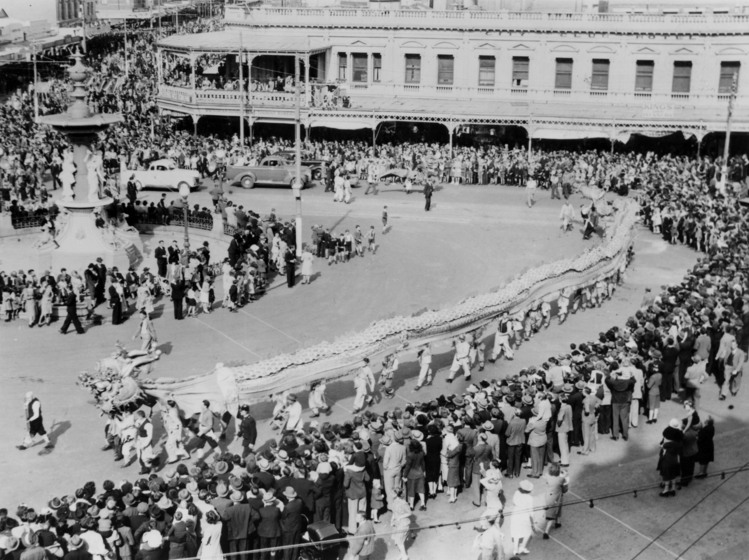 Black and white photograph of a long Chinese ceremonial dragon in a procession with crowds lining the street, taken from a high vantage point.