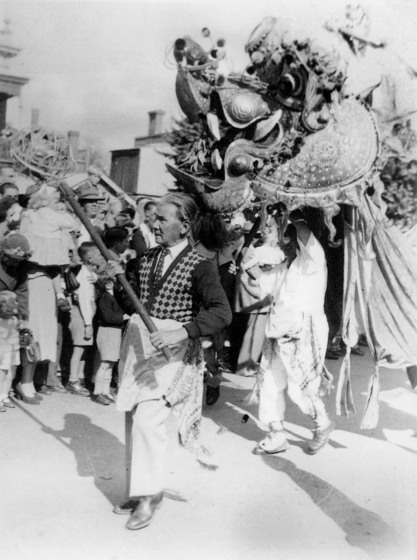 Black and white photograph of a person supporting a large processional dragon's head with another holding a ball on a pole.