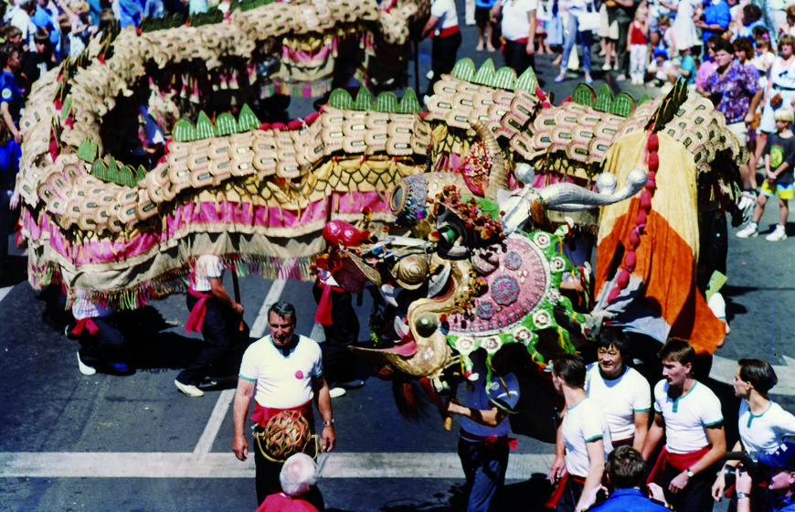 Colour photograph of a long Chinese ceremonial dragon in a procession with five men in white T-shirts and red sashes in the foreground.