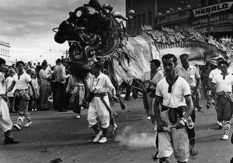 Black and white photograph of a group of men in white T-shirts leading a large ceremonial dragon in a street procession.