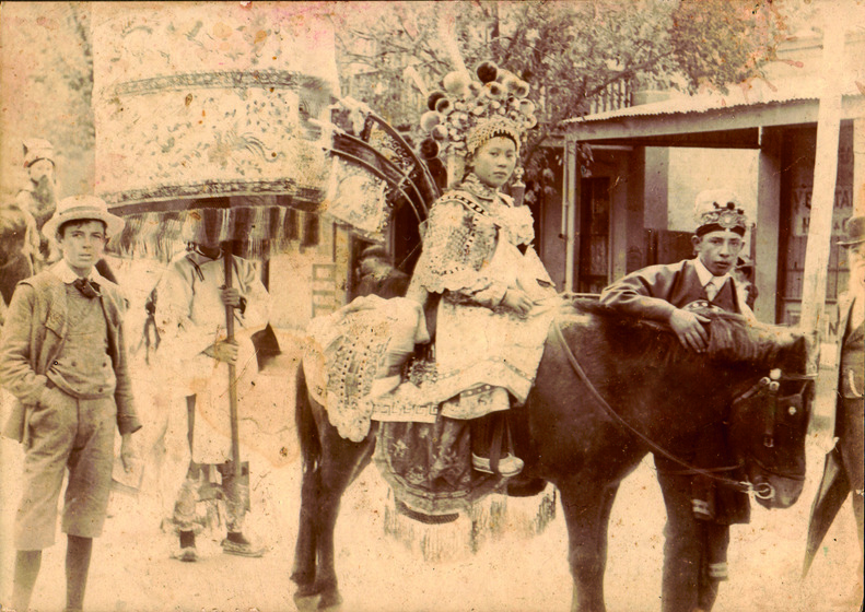 Sepia photograph of a young Asian woman in an elaborate costume and headdress riding a pony followed by a person in costume carrying a large decorative lantern.