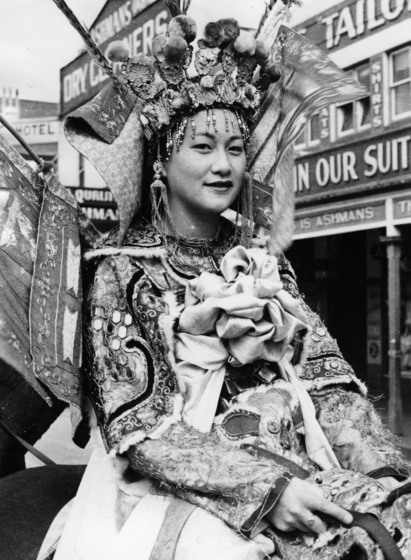 Black and white close-up photograph of a young Asian woman in an elaborate costume and headdress, shop signage behind her.