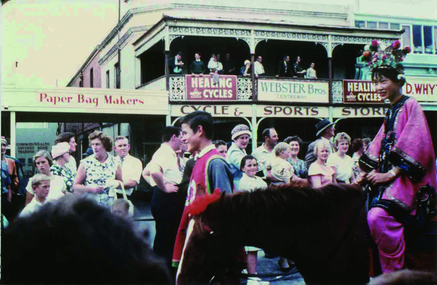 Colour photograph of a young Asian woman in a pink costume with a headdress riding a chestnut pony being led by a boy. Crowd on side of street and on first floor verandah look on.