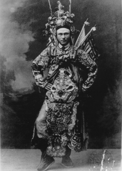 Black and white full-length studio photo portrait of a man in an elaborate costume with a dragon on the front, a headdress and flags attached behind.