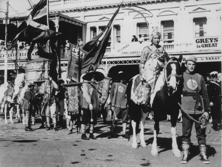 Black and white photograph of an Asian man in an elaborate costume and headdress riding a horse led by a young man in costume and leading a street procession. People look on from first floor balconies and window.