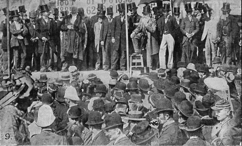 Black and white newspaper clipping of a photograph of a row of men in Victorian top hats on a stage with a crowd of people in bowler hats and pith helmets below.