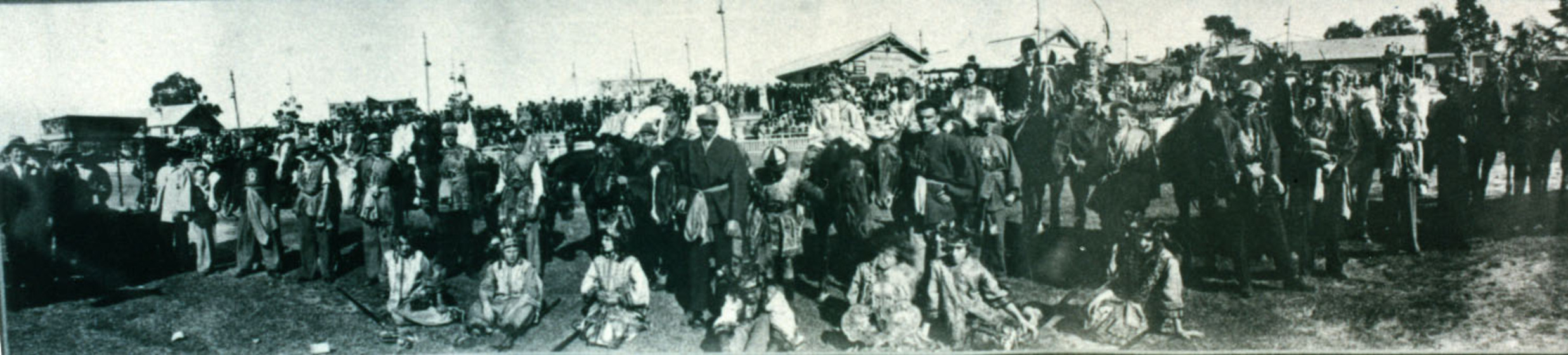 Wide black and white photograph of a row of costumed people, some on horseback.