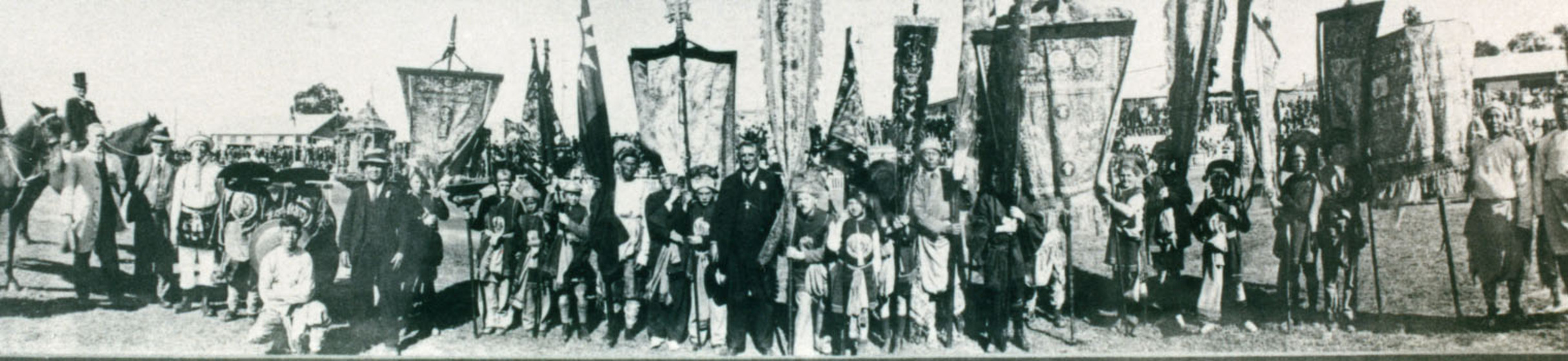 Wide black and white photograph of a row of costumed people some with banners.
