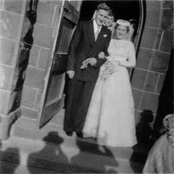 Black and white photograph of a bride and groom at a church door with the photographer's shadow in the foreground.