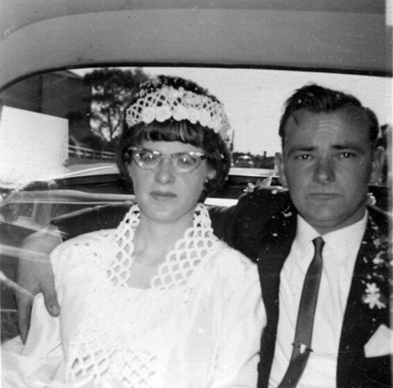 Black and white photograph of a bride and groom in the back of a car. The groom has his arm around the bride. Neither are smiling.