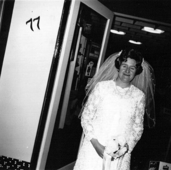 Black and white photograph of a bride standing in a doorway with the number 77 on the wall beside her.