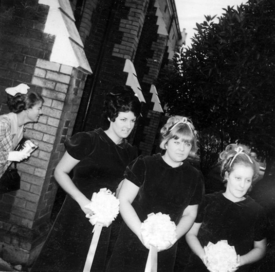 Black and white photograph of three bridesmaids in dark dresses standing in a row outside a church.