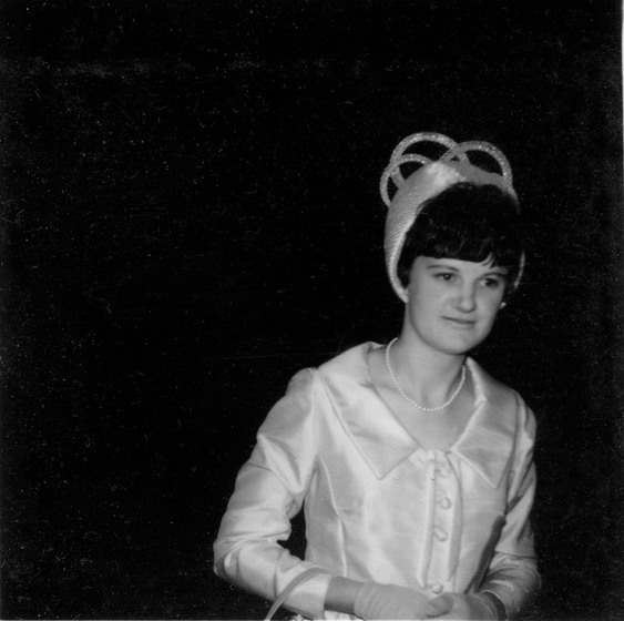 Black and white photograph of a woman in a pale 1960s dress and pill box hat with loops.
