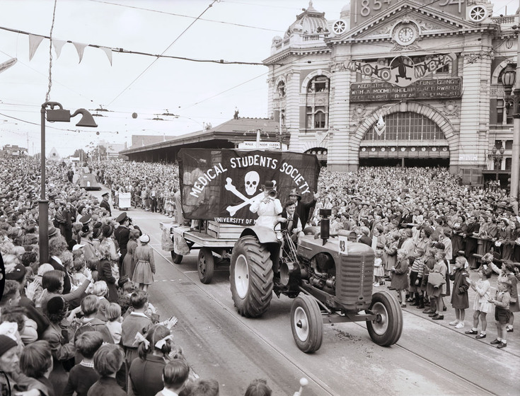 Black and white photograph of a street parade with a float drawn by a tractor and carrying the banner 'Medical Students Society'. A large crowd of onlookers.