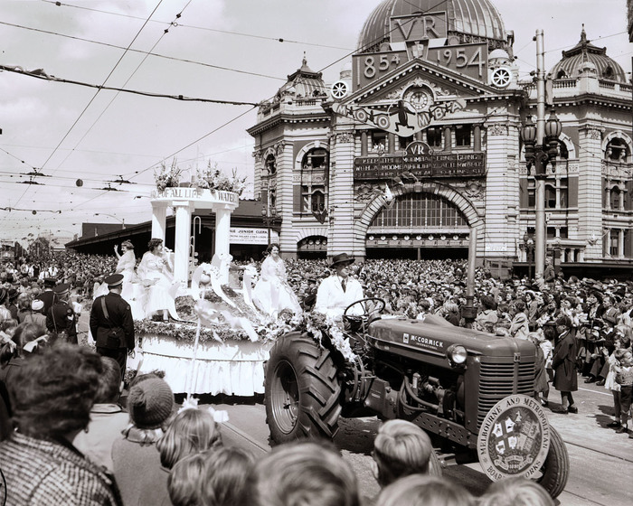 Black and white photograph of a street parade with a white circular float drawn by a tractor and carrying four women in white gowns. A large crowd of onlookers.