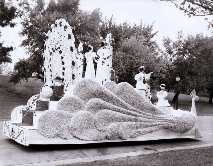 Black and white photograph of a parade float in the shape of a peacock with four women in white gowns on it.