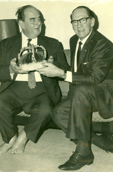 Black and white photograph of two seated men in suits holding a crown.