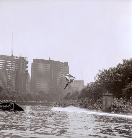 Black and white photograph of a water skier with a kite airborne over a river. Crowd on riverbank. Multi-storey buildings in distance.