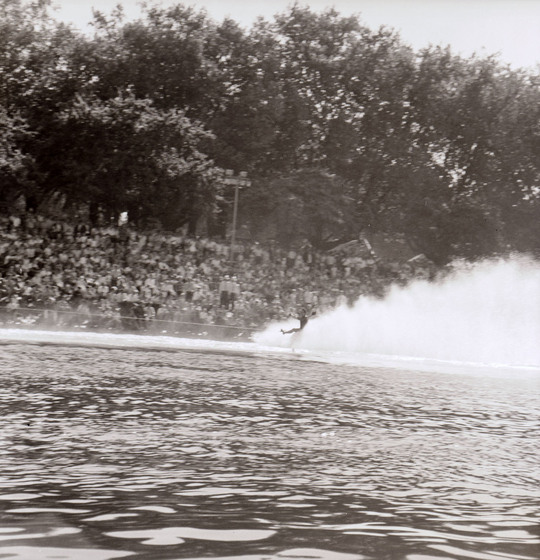 Black and white photograph of a man water skiing one legged river. Crowd on riverbank.
