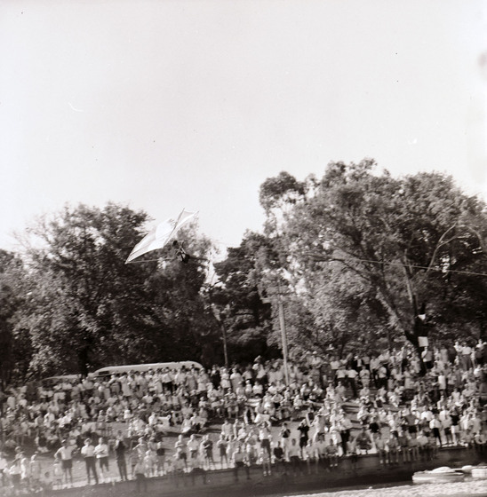 Black and white photograph of a water skier with a kite airborne over a river. Crowd on riverbank.