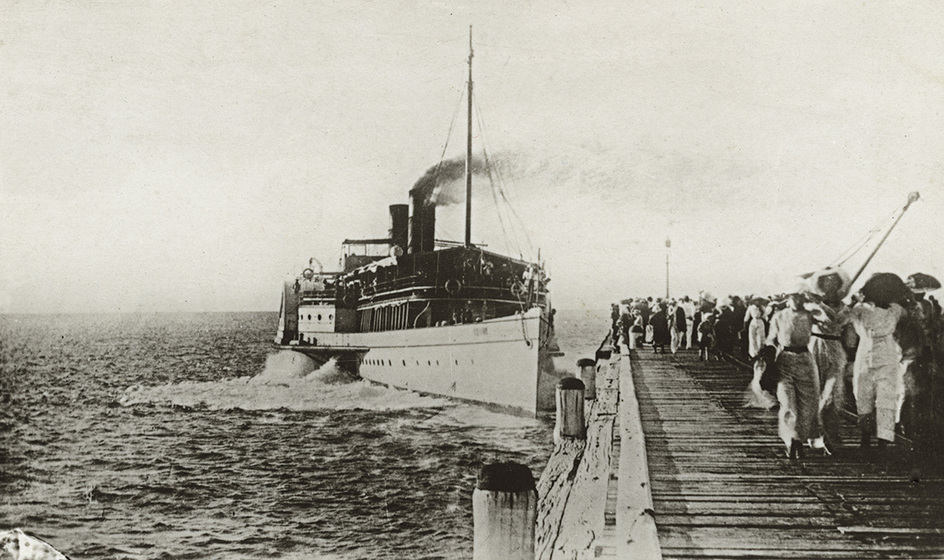Black and white photographic postcard of a paddle steamer approaching a crowd on a wooden pier.