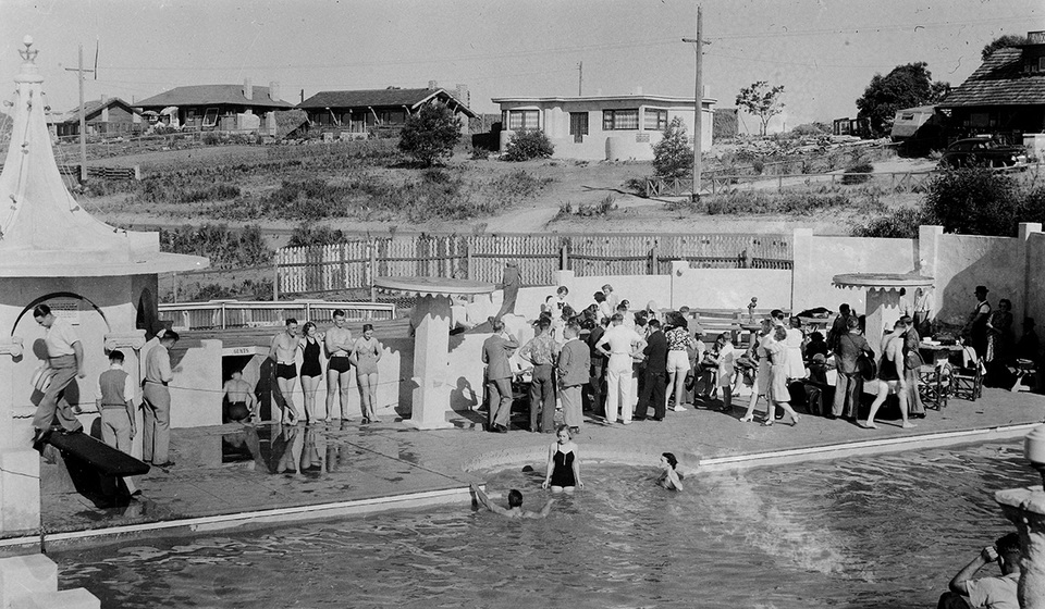 Black and white photographic postcard of a crowd beside a swimming pool. Houses in middle distance.
