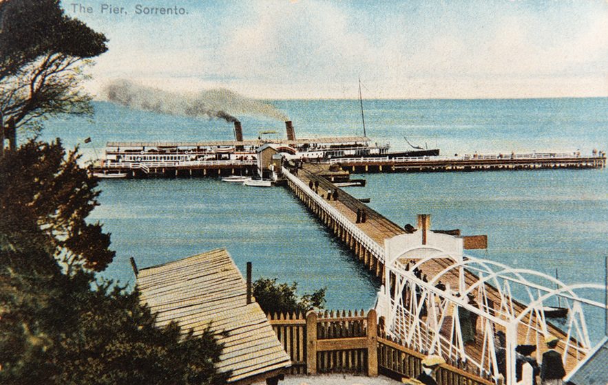 Tinted black and white photographic postcard of a long pier with a paddle steamer.