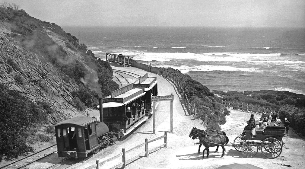 Black and white photographic postcard of a small steam locomotive pulling two open tramcars on a coastal track. Two horses pulling a group of people in an open carriage in the foreground. Surf breaking in the distance.