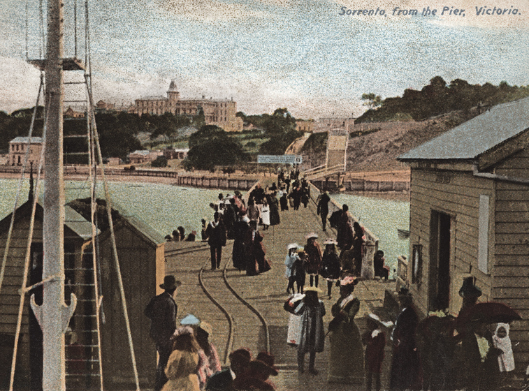 Tinted black and white photographic postcard of a crowd on a pier looking to a large building on the shore.