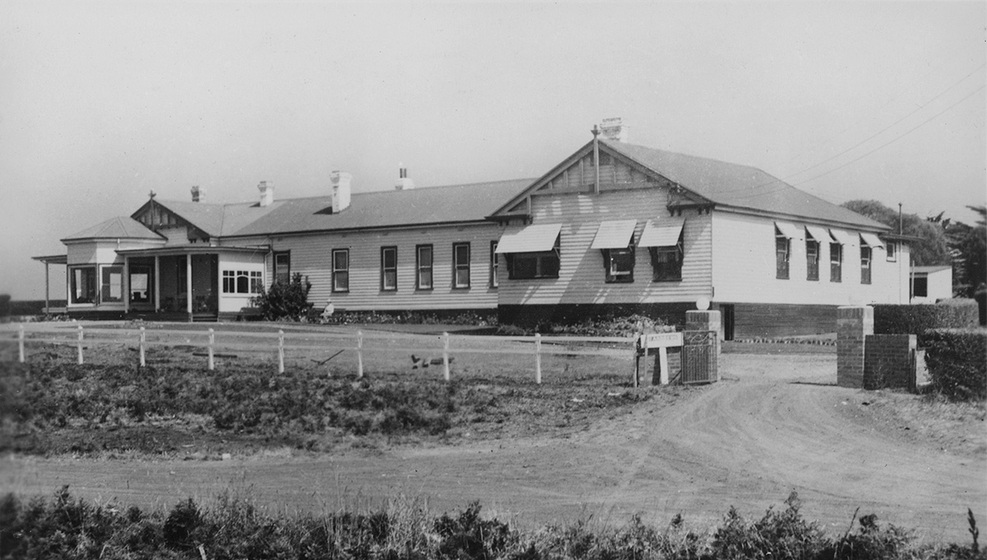 Black and white photographic postcard of a large single storey white weatherboard building with awnings on the windows.