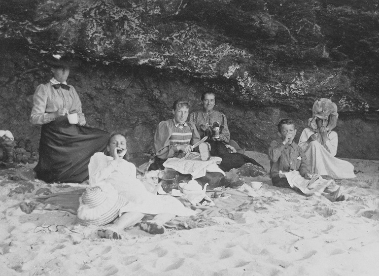 Black and white photograph of three women, two girls and a boy in Edwardian dress picnicking on a beach.