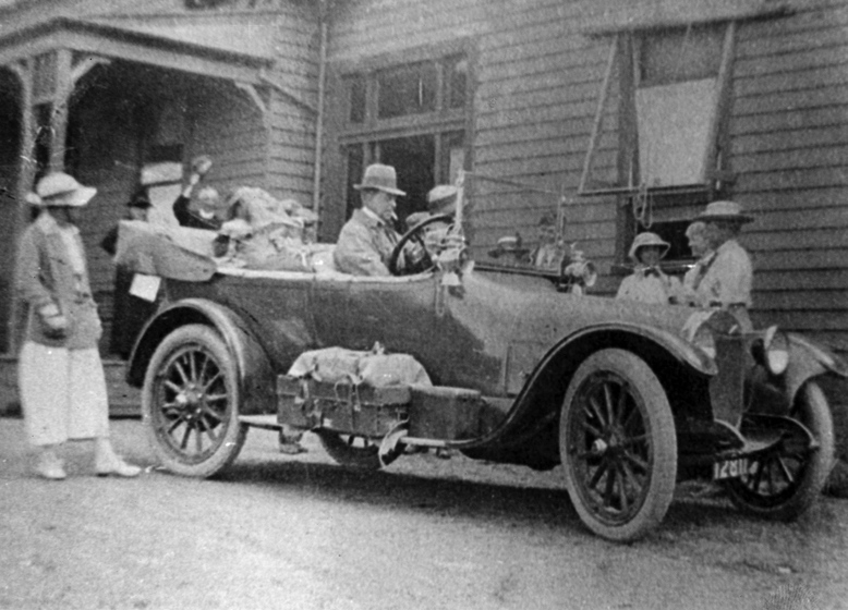 Black and white photograph of a group standing around an early open automobile with a driver outside a weatherboard building.
