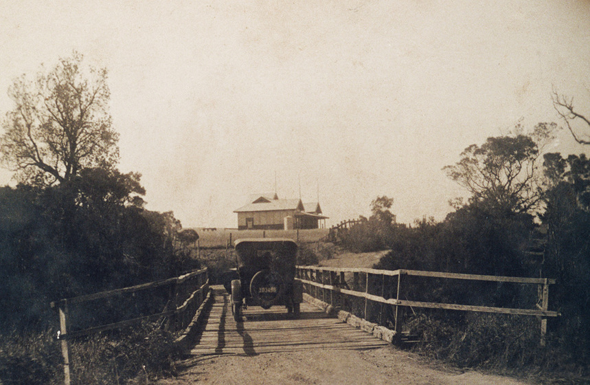 Sepia photograph of early car on wooden bridge with a single storey wooden building on the hill above.