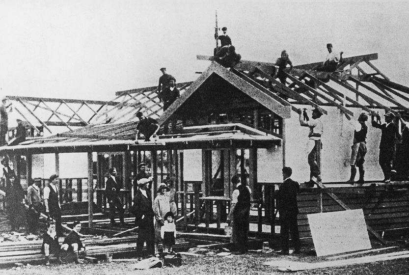 Black and white photograph of a single storey wooden structure with a gable roof under construction. Men, women and children watch people building.