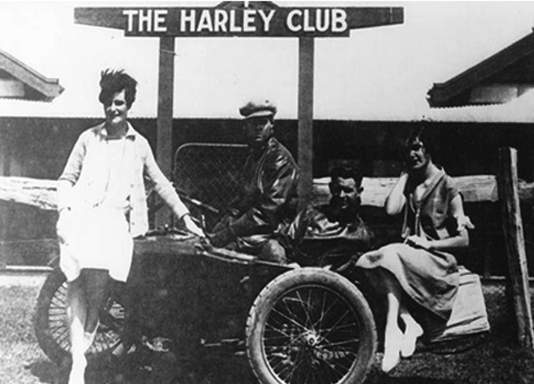 Black and white photograph of two men in leathers on a motorbike with a side car. Two women in 1930s clothes lean on the bike. Sign above them reads, 'The Harley Club.'
