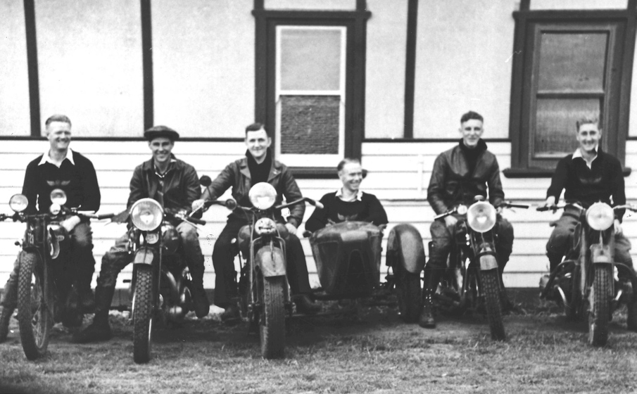 Black and white photograph of a row of five stationary motorbikes with riders in front of the wall of a wooden building.