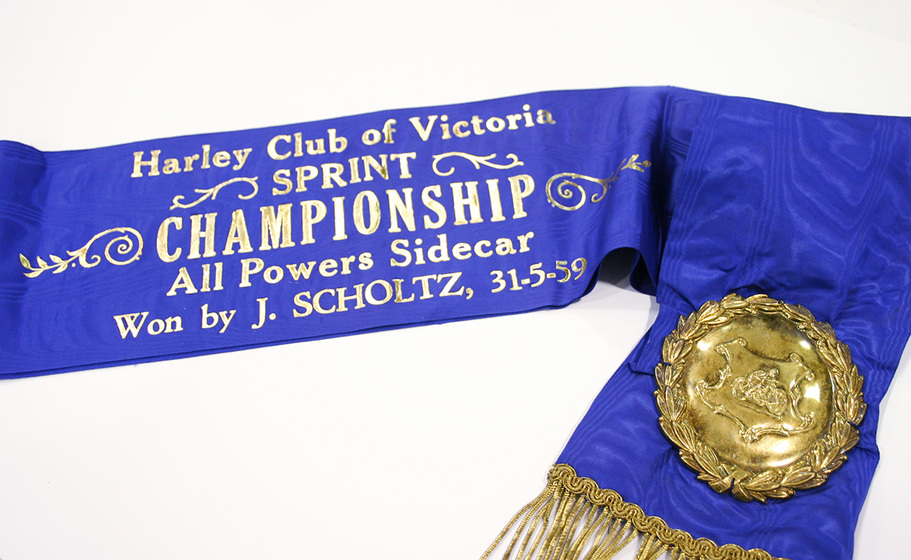 Blue sash with embossed gold-coloured medallion and fringe and the inscription in gold, 'Harley Club of Victoria Sprint Championship All Powers Sidecar Won by J. Scholtz 31-5-59.'
