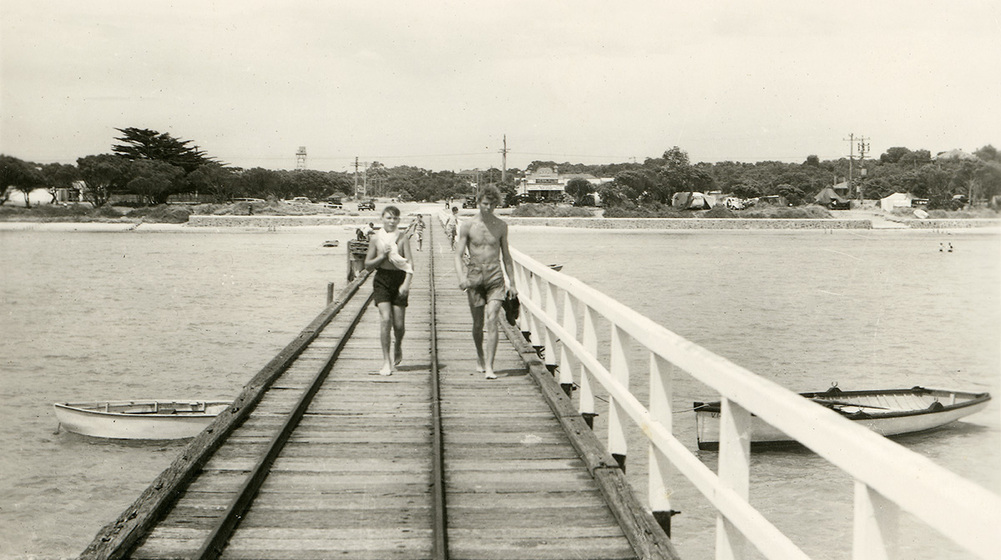 Black and white photograph looking along a wooden from the sea to the shore. Two boys in shorts walking toward the camera.