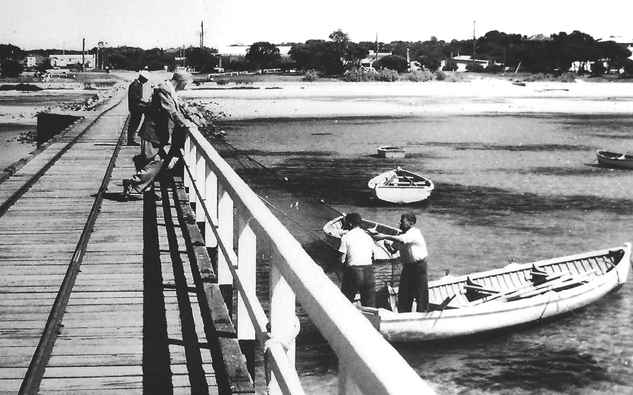 Black and white photograph looking along a wooden from the sea to the shore. Two men on the pier watch two men in a boat with a net.