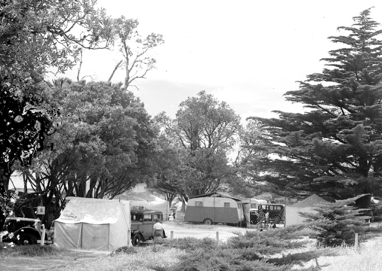Black and white photographic postcard of cars, tents and caravans amongst trees.