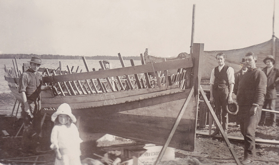 Black and white photograph of six men an small girl standing around a wooden boat under construction on a foreshore.
