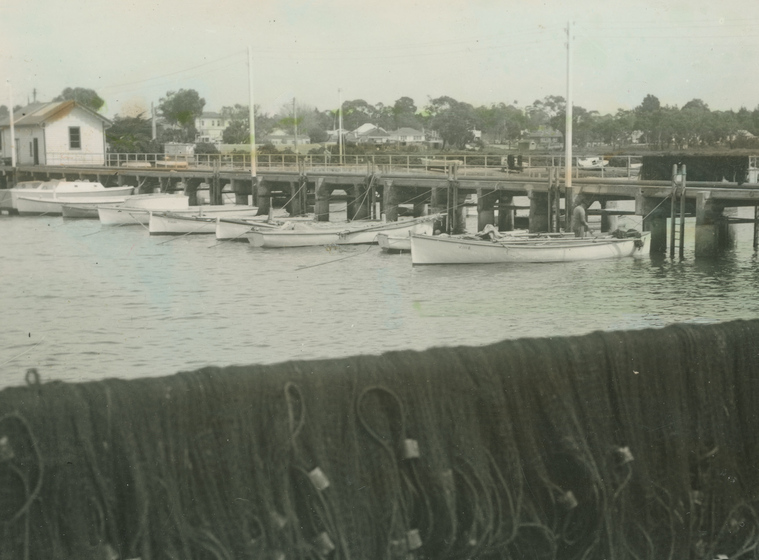 Black and white photographic postcard of a row of sailing boats moored at a wooden pier. Fishing nets in foreground.
