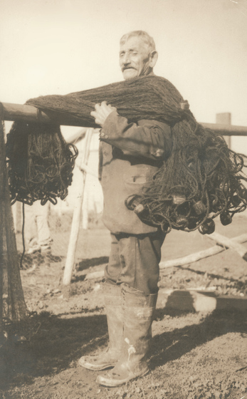 Sepia photograph of old man with moustache in gumboots carrying a fishing net over one shoulder.