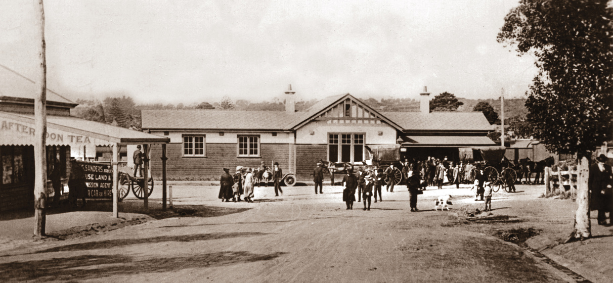 Black and white photograph of a street scene with a crowd and horse drawn carriages outside a low wooden building.