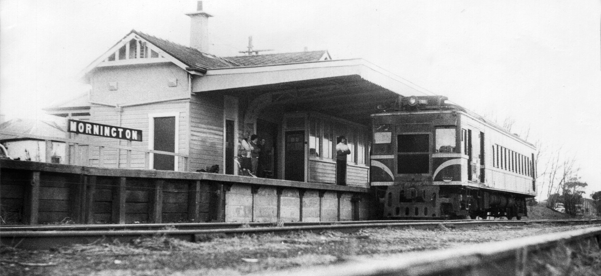Black and white photograph of a train engine at a platform at a wooden station building with a sign saying, 'Mornington.'
