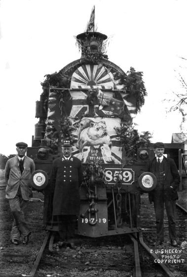 Black and white photograph of a three men, one in a uniform, standing on train tracks in front of a steam train engine decorated with a picture of a British bulldog and other British empire insignia.