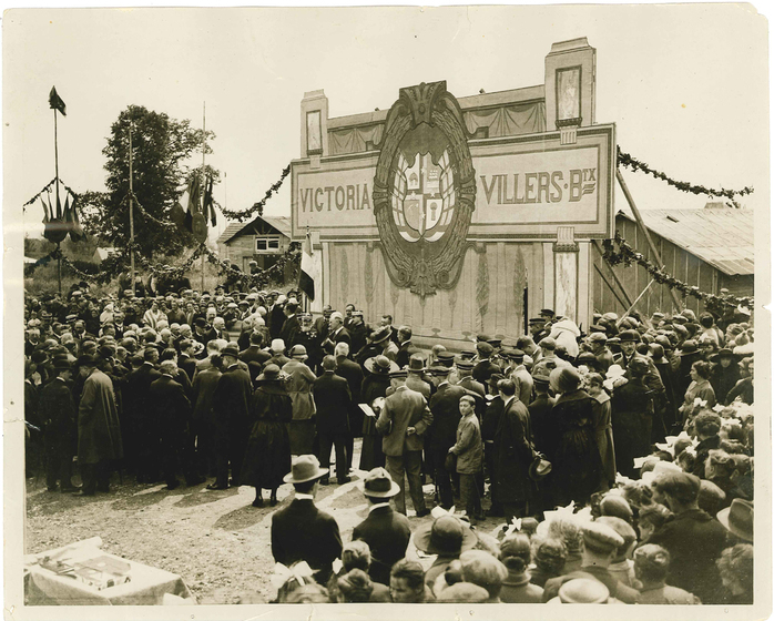 Black and white photograph of a crowd in 1920s clothing listening to a speaker in front of a backdrop with a coat of arms painted on it and the words, 'Victoria Villers'.