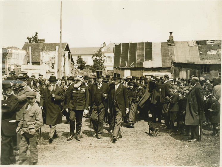 Black and white photograph of two men in top hats and morning coats striding through a rural town surrounded by a crowd.